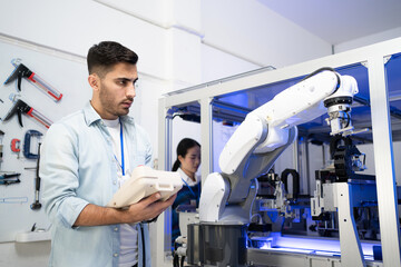 Fototapeta na wymiar Young Caucasian male industrial engineer in a blue shirt standing controlling an automation robotic arm machine for multiple industrial applications with a robot controller in an industrial factory.