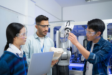 Caucasian male engineer standing hold a robot controller next to Asian female apprentice holding a laptop, demonstrating the work of robotic machine to young Asian male CEO who smile touching a robot. - obrazy, fototapety, plakaty