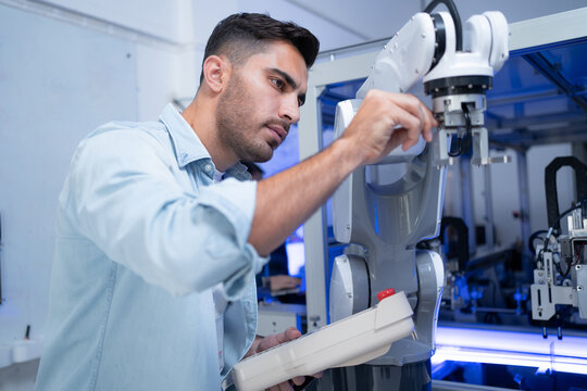 Young Caucasian male engineer in blue shirt touching and checking automation robotic arm machine with a robotic machine controller in hand while the inspection of the machine in an industrial factory.