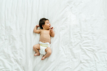 Child Care Concept. Above top high view portrait of cute little caucasian newborn baby wearing diaper lying on the white blanket sheets at home with finger in mouth. Infant girl child resting at home. - obrazy, fototapety, plakaty