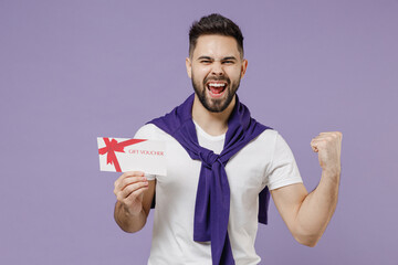 Satisfied cheerful young brunet man 20s wears white t-shirt purple shirt hold gift certificate coupon voucher card for store doing winner gesture isolated on pastel violet background studio portrait - obrazy, fototapety, plakaty