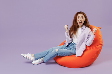 Full size body length young redhead woman 20s wears white T-shirt violet jacket sit in bag chair do winner gesture clench fists say yes isolated on pastel purple color wall background studio portrait.