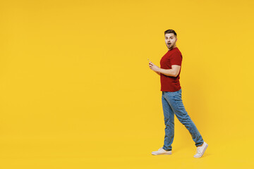 Fototapeta na wymiar Full length side view happy smiling young man in red t-shirt casual clothes using mobile cell phone walk going isolated on plain yellow color wall background studio portrait. People lifestyle concept.
