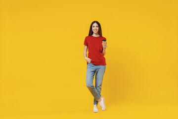 Fototapeta na wymiar Full size body length young brunette woman 20s wears basic red t-shirt hold takeaway delivery craft paper brown cup coffee to go isolated on yellow background studio portrait. People emotions concept.