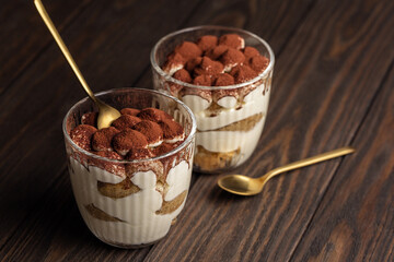 Tiramisu in glass on wooden table, traditional coffee flavored Italian dessert made of savoyardi cookies, mascarpone cream and cocoa powder.