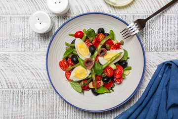 French Nicoise salad with anchovy, green beans, tomatoes, eggs, black olives, capers on white plate and white wooden background. Healthy food, no carb diet concept.