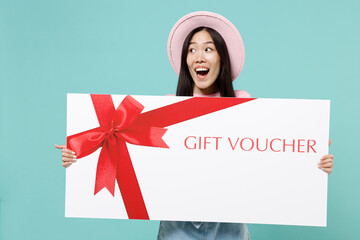 Overjoyed young brunette asian woman 20s wears pink clothes hold huge big great size gift certificate coupon voucher card for store look aside isolated on pastel blue color background studio portrait