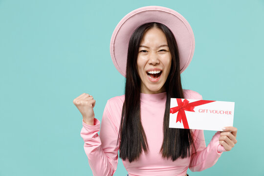 Overjoyed young brunette asian woman 20s wears pink clothes hold gift certificate coupon voucher card for store celebrate clench fists say yes isolated on pastel blue color background studio portrait