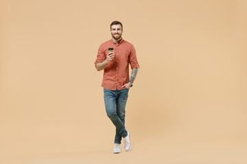 Fototapeta na wymiar Full size body length happy tatooed young brunet man 20s short haircut wear apricot shirthold takeaway delivery craft paper brown cup coffee to go isolated on pastel orange background studio portrait