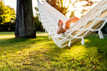 Cute little feet of barefoot child sticking out of hammock in backyard garden at sunset. Child summer holidays healthy lifestyle, outdoors leisure activity. Vacation at home, slow living, gadget detox