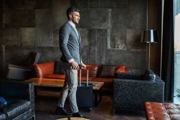 A businessman carrying his luggage in the lobby of a modern hotel. He is on a business trip and is...