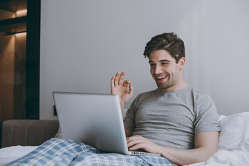 Young man in pajamas grey t-shirt lying sit in bed talk by video call use work laptop pc computer show okay gesture rest relax at home indoors bedroom. Good mood sleeping night morning bedtime concept