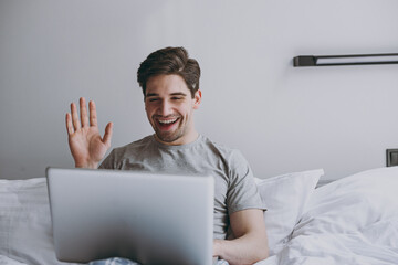 Young man in pajamas grey t-shirt lying sit in bed talk by video call use work on laptop pc computer waving hand rest relax at home indoors bedroom. Good mood sleeping night morning bedtime concept