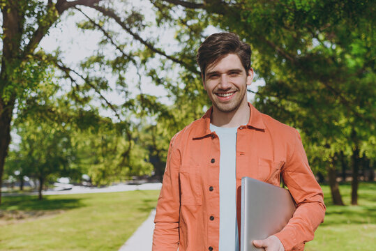 Close up young man in orange jacket blue t-shirt walk hold closed lapop pc computer look aside rest relax in spring green city park go down alley sunshine lawn outdoor on nature Urban leisure concept