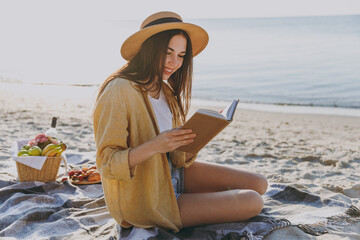 Full size young happy traveler tourist woman in straw hat shirt summer clothes reading book sit on plaid have picnic outdoors on sea sand beach background People vacation lifestyle journey concept
