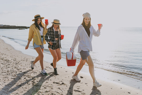 Full Length Female Friends Young Women In Straw Hat Summer Clothes Hang Out Together Carry Food In Picnic Refrigerator Cups Walking Go Outdoor On Sea Beach Background People Vacation Journey Concept.