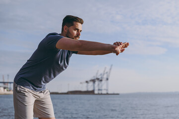 Young strong sporty athletic toned fit sportsman man in sports clothes warm up training do hand exercise forward tilting at sunrise sun over sea beach outdoor on pier seaside in summer day morning.