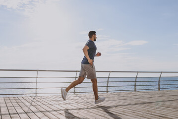 Full body side view young strong sporty athletic fit sportsman man in sports clothes warm up training run jogging at sunrise sun over sea beach outdoor on pier seaside in summer day cloudy morning