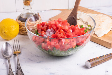 Cooking Mediterranean vegetarian salad with tomatoes, onions and herbs, mixing chopped vegetables and herbs in a glass bowl on a light table, background