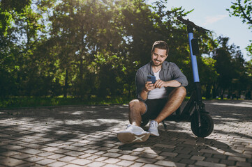 Fototapeta premium Full length bottom view young man in blue shirt sit near riding scooter hold mobile cell phone go down alley rest in spring green city sunshine park outdoors on nature Urban leisure lifestyle concept.