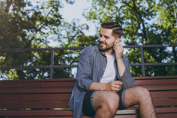 Smiling young man 20s wearing blue shirt shorts earphones sit on bench use mobile cell phone listen...