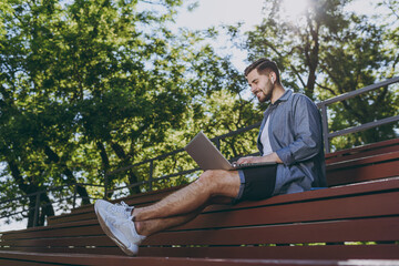 Bottom view full body young fun man in blue shirt shorts earphones sit on bench work online laptop pc computer waving hand rest relax in spring green city park outdoor on nature Urban leisure concept.