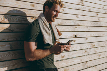 Obraz premium Young strong sporty athletic fit sportsman man wear sports clothes towel warm up training use mobile cell phone surfing internet leaning on wooden wall sea beach outdoor seaside in summer day morning.