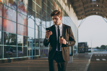 Traveler businessman young man in black suit going walk outside at international airport terminal use mobile phone book taxi order hotel hold paper cup coffee to go Air flight business trip concept