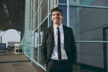 Bottom view happy satisfied young traveler businessman man in black classic tie suit standing outside at international airport terminal look aside People air flight business trip lifestyle concept.