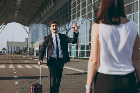 Young happy traveler businessman man 20s wearing black dinner suit walk go outside at international airport terminal with suitcase valise waving hand meet woman wife Air flight business trip concept