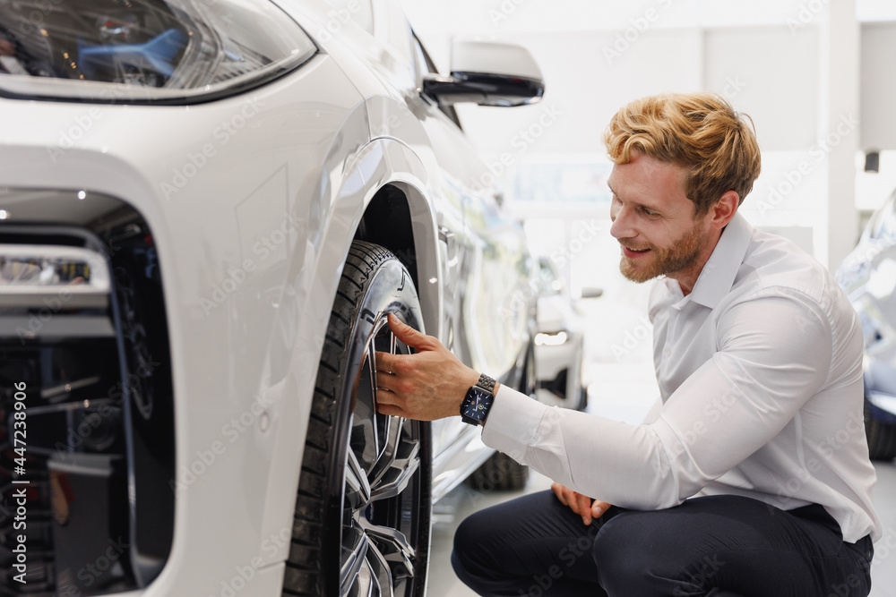 Wall mural Man happy smiling customer male buyer client wear white shirt touch tire wheel disc choose auto want buy new car automobile in showroom salon vehicle dealership store motor show indoor Sales concept