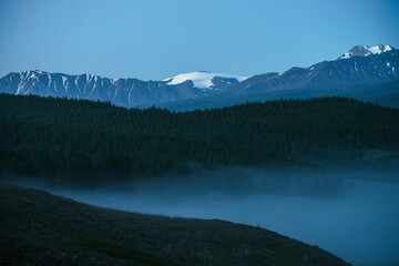 Naklejka na ściany i meble Atmospheric mountains landscape with dense fog and great snow mountain top under twilight sky. Alpine scenery with big snowy mountains over thick fog in night. High snow pinnacle above clouds in dusk.