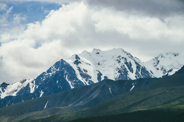 Awesome mountains landscape with high snowy mountain range among white clouds in blue sky. Atmospheric highland scenery with snow-white big mountain ridge in overcast weather. Wonderful snowy pinnacle