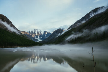 Atmospheric alpine landscape with snowy mountains in golden sunlight reflected on mirror mountain lake in fog among low clouds. Scenic highland scenery with low clouds on rocks and green mirror lake.
