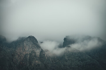 Dark atmospheric mountain landscape with sharp rocks in low clouds. Dark rocks in gray overcast weather. Gloomy mountain scenery with gray low clouds on rough crags. Minimalism with rocky mountains.