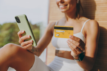 Cropped up photo young sportswoman woman in white sportswear earphones listen music use mobile phone credit bank card shop online order delivery book tour pay gym membership sit on windowsill indoor.