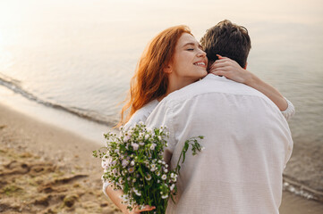 Satisfied young lovely couple two family man woman in white clothes hug each other hold flowers rest together at sunrise over sea ocean sand beach outdoor exotic seaside in summer day sunset evening.