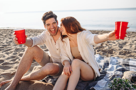 Young happy smiling couple family two man woman in white clothes hug sitting on sand plaid have picnic drink hold red cups rest together at sunrise over sea beach ocean outdoor seaside in summer day.