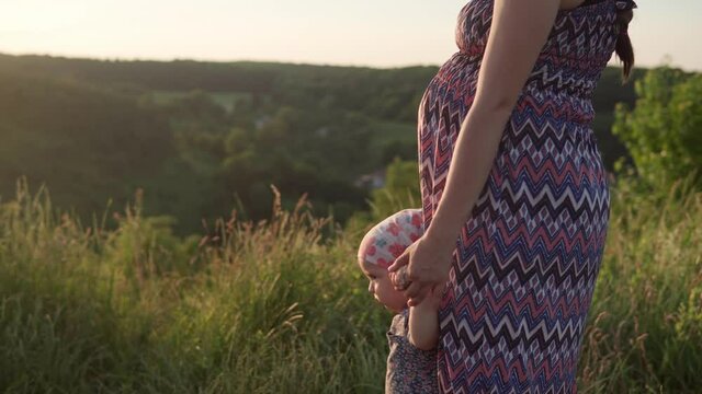 cute young pregnant woman in long dress hat with toddler girl on top of mountain before sunset. people in park. happy mom and baby daughter holding hands. childhood, nature, lifestyle, summer concept