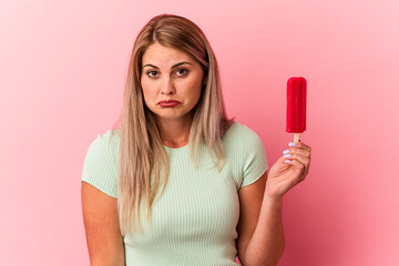 Young russian woman holding an ice cream isolated on pink background shrugs shoulders and open eyes...