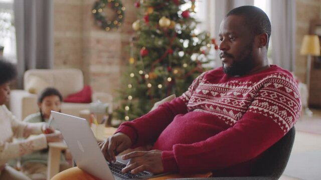 Afro-American man sitting at home and using laptop while wife and daughter drawing near Christmas tree in background