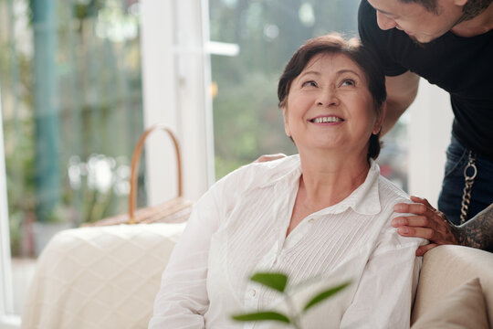 Asian matue woman sitting on sofa smiling and talking to her elder son at home