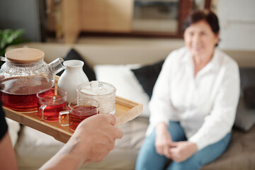 Close-up of son holding tray with teapot and cups and serving it for his mother while she sitting on sofa in the room