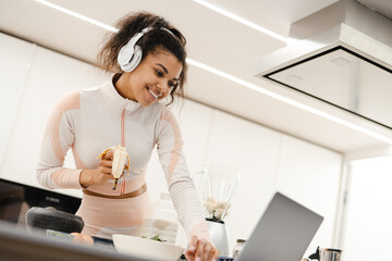 Black woman in headphones using laptop while making smoothie at home