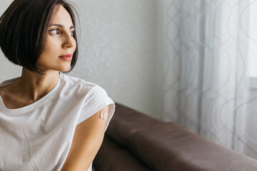 Naklejka premium Portrait of smiling woman after getting a vaccine. Female holding down her white shirt sleeve and showing her arm with bandage after receiving vaccination. Concept of recommended inoculation