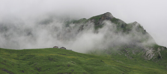 Farm houses, green meadow and mountain peak reaching out of fog and clouds.