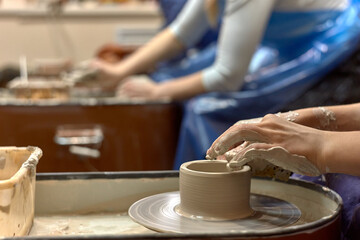 Hands of a ceramist while she is working on a potter's wheel against the background of other people working with clay