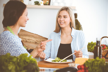 Two women friends taste new recipes for delicious fresh salads while sitting at the kitchen table. Vegetarian cooking concept