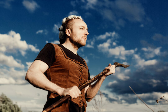 A young man with an interesting depicts a Viking with an ax in an unreliable stylized modern costume looking into the distance against a blue sky with clouds.