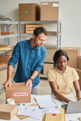 Vertical portrait of two young people using laptop in warehouse while managing small business